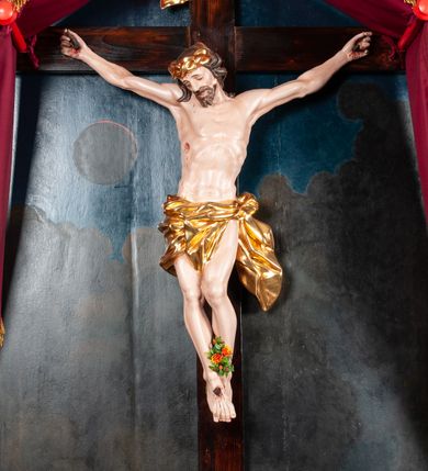Zdjęcie nr 2: Rzeźbiony krucyfiks, zawieszony na prostym krzyżu na tle obrazu z przedstawieniem zaćmienia słońca w górnej części i widokiem Jerozolimy za wzgórzami w dolnej, w niszy zamkniętej od góry daszkiem dwuspadowym. W centrum rzeźbiona figura Chrystusa Ukrzyżowanego, z rozłożonymi, nieznacznie ugiętymi rękami przybitymi gwoździami, ze stopami złożonymi razem, prawa na lewej, przybitymi jednym gwoździem; lewe kolano ugięte, wysunięte przed prawe. Prawe biodro lekko wygięte, ciało tworzy mało wydatny łuk. Na torsie widoczna rana w prawym boku. Głowa opada na prawe ramię, twarz owalna, oczy zamknięte, brwi lekko uniesione, nos prosty, widoczne krótkie wąsy i dzieląca się w dwa pukle broda. Kręcone włosy opadają na kark i prawe ramię. Zarost i włosy jasnobrązowe. Na głowie masywna, złocona korona cierniowa. Biodra przewiązane złoconym, krótkim perizonium związanym na lewym biodrze, układającym się w drobne, poprzeczne fałdy. Koniec perizonium powiewa przy lewym udzie. Chrystus wisi na drewnianym, ciemnobrązowym krzyżu o długiej belce pionowej. W jej górnej części titulus z napisem „INRI”. 