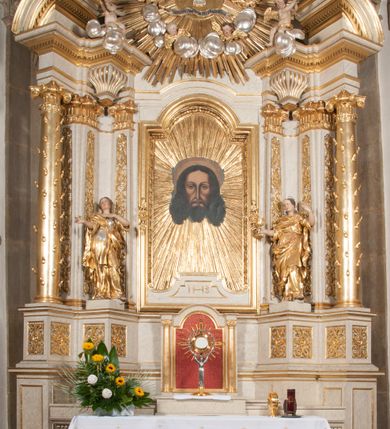 Zdjęcie nr 2: Ołtarz architektoniczny, przyścienny, jednoosiowy, jednokondygnacyjny z mensą. Mensa prosta, kamienna; antepedium płaskorzeźbione ze sceną Zesłania Ducha Świętego. Kompozycja jest niemal symetryczna, postacie apostołów oraz dwie Marie siedzą lub stoją poniżej rozchodzących się promieni glorii. Nastawa ołtarzowa ustawiona na wysokim, zdwojonym cokole, którego szersza oś środkowa jest flankowana parami cokołów ustawionych skośnie. Pierwsza kondygnacja również ustawiona na cokole, którego pola wypełniają płyciny z płaskorzeźbioną, symetryczną dekoracją z ceownicami wokół kwiatu róży. Na środku, na mensie prostopadłościenne tabernakulum na cokole. Na jego ścianie frontowej wypełnione czerwonym kolorem drzwi w kształcie stojącego prostokąta zamkniętego od góry łukiem nadwieszonym, flankowane kolumienkami jońskimi. 
W polu środkowym pierwszej kondygnacji ołtarza obraz „Oblicze Chrystusa” (XVII w.) w profilowanej, prostokątnej ramie z uskokiem i półkolistym zamknięciem od góry. Pole flankowane skośnie ustawionymi kolumnami korynckimi na tle pilastrów, wspierającymi wyłamane nad nimi belkowanie i fragmenty przerwanego przyczółka. Trzony kolumn ozdobione plastycznymi płomieniami. Na pilastrach płyciny z wypełnieniem z płaskorzeźbionych ornamentów o motywach regencyjnych. Na tle pilastrów, po obu stronach środkowego obrazu, rzeźbione figury św. Heleny i św. Barbary w obficie drapowanych szatach. 
Zwieńczenie ołtarza w formie glorii promienistej z gołębicą Ducha Świętego pośrodku. Poniżej niej, na banderoli inskrypcja „EMITTE  SPIRITUM TUUM ET CREABUNTUR”. Przedstawienie ujęte otokiem utworzonym z obłoków i uskrzydlonych główek. Po bokach, na częściach przerwanego belkowania, na obłokach siedzą putta. Są zwrócone do siebie i spoglądają lekko w dół. Mają uniesione ręce i są przepasane złotymi szarfami. 
Struktura ołtarza dekorowana imitacją marmoryzacji za pomocą polichromii na kolor jasnoszary z ciemniejszymi żyłkami; profile, ornamenty, gloria, szaty świętych złocone; obłoki, gołębica, elementy strojów, fragmenty antepedium srebrzone; polichromia w partiach ciała postaci naturalistyczna. 