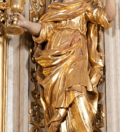 Zdjęcie nr 7: Figura pełna, na cokole, przedstawiająca św. Barbarę. Święta w pozie przypominającej literę „S”, w kontrapoście z ciężarem na prawej nodze, z lewą odstawioną do boku. Obie ręce zgięte w łokciach, w prawej trzyma kielich, lewą unosi do góry w delikatnym geście. Głowa przechylona na prawo, twarz o łagodnym wyrazie, okrągła, z wysokim czołem, małymi, przymkniętymi oczami, niewielkim nosem i wąskimi ustami; brązowe włosy zaczesane do tyłu, okryte chustką. Święta jest ubrana w długą, przepasaną suknię, która podkreśla kształt ciała, przylegając do niego drobnymi fałdami; rękawy sukni szerokie, sięgające łokci. Barbara na suknię ma zarzucony płaszcz, który przykrywa dekolt, owija się wokół prawej ręki i jest podczepiony do paska na lewym biodrze. 
Polichromia w partiach ciała naturalistyczna, szaty i atrybut złocone, chustka srebrzona. 