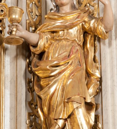 Zdjęcie nr 6: Figura pełna, na cokole, przedstawiająca św. Barbarę. Święta w pozie przypominającej literę „S”, w kontrapoście z ciężarem na prawej nodze, z lewą odstawioną do boku. Obie ręce zgięte w łokciach, w prawej trzyma kielich, lewą unosi do góry w delikatnym geście. Głowa przechylona na prawo, twarz o łagodnym wyrazie, okrągła, z wysokim czołem, małymi, przymkniętymi oczami, niewielkim nosem i wąskimi ustami; brązowe włosy zaczesane do tyłu, okryte chustką. Święta jest ubrana w długą, przepasaną suknię, która podkreśla kształt ciała, przylegając do niego drobnymi fałdami; rękawy sukni szerokie, sięgające łokci. Barbara na suknię ma zarzucony płaszcz, który przykrywa dekolt, owija się wokół prawej ręki i jest podczepiony do paska na lewym biodrze. 
Polichromia w partiach ciała naturalistyczna, szaty i atrybut złocone, chustka srebrzona. 