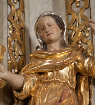 Zdjęcie nr 4: Figura pełna, na cokole, przedstawiająca św. Barbarę. Święta w pozie przypominającej literę „S”, w kontrapoście z ciężarem na prawej nodze, z lewą odstawioną do boku. Obie ręce zgięte w łokciach, w prawej trzyma kielich, lewą unosi do góry w delikatnym geście. Głowa przechylona na prawo, twarz o łagodnym wyrazie, okrągła, z wysokim czołem, małymi, przymkniętymi oczami, niewielkim nosem i wąskimi ustami; brązowe włosy zaczesane do tyłu, okryte chustką. Święta jest ubrana w długą, przepasaną suknię, która podkreśla kształt ciała, przylegając do niego drobnymi fałdami; rękawy sukni szerokie, sięgające łokci. Barbara na suknię ma zarzucony płaszcz, który przykrywa dekolt, owija się wokół prawej ręki i jest podczepiony do paska na lewym biodrze. 
Polichromia w partiach ciała naturalistyczna, szaty i atrybut złocone, chustka srebrzona. 