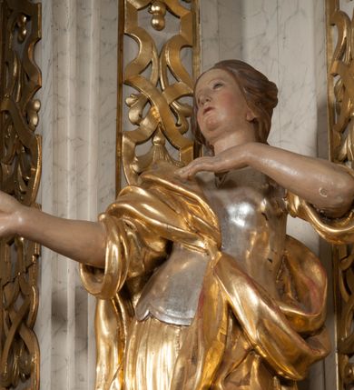 Zdjęcie nr 3: Figura pełna na cokole przedstawiająca św. Helenę. Święta w pozie przypominającej literę „S”, w kontrapoście z ciężarem na prawej nodze, z lewą odstawioną do boku. Obie ręce zgięte w łokciach, prawa wyciągnięta przed siebie z palcami ułożonymi jakby coś trzymały, lewą unosi do piersi. Głowa skręcona lekko w prawo, twarz o łagodnym wyrazie, owalna, z wysokim czołem, małymi oczami, niewielkim nosem i wąskimi ustami; brązowe włosy zaczesane do tyłu opadają na plecy. Święta jest ubrana w długą suknię z gorsetem, której dolna część przylega do ciała drobnymi fałdami. Na suknię ma zarzucony płaszcz, który przechodzi skośnie przez pierś, nad prawą ręką i wokół lewego biodra. 
Polichromia w partiach ciała naturalistyczna, gorset srebrzony, suknia i płaszcz złocone. 