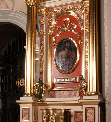 Zdjęcie nr 2: Ołtarz architektoniczny, przyścienny, jednokondygnacyjny, jednoosiowy, w formie podwójnej aediculi. Mensa prostopadłościenna, z płyciną w kształcie leżącego prostokąta wypełnionego marmoryzacją i obwiedzionego profilowaną ramą. Na mensie tabernakulum na planie zbliżonym do owalu, o wklęsło-wypukłej linii ścian. Części boczne na cokole, w środkowej drzwiczki w kształcie stojącego prostokąta zamkniętego łukiem nadwieszonym z symetryczną dekoracją składającą się z kielicha z hostią otoczoną glorią promienistą na tle kłosów zbóż oraz kiści winogron. Na bocznych ściankach dekoracja z ornamentu rocaille&#039;owego. Tabernakulum od góry zamknięte profilowanym gzymsem. 
Nastawa na wysokim, trójdzielnym cokole dekorowanym prostokątnymi płycinami. W polu głównym rama w kształcie stojącego prostokąta zamkniętego od góry łukiem wklęsło-wypukłym z uskokiem. W niej obraz z przedstawieniem Ecce Homo w owalnej ramie poniżej kokardy uwitej ze złotej wstęgi, i powyżej tabliczki z napisem „O MÓJ JEZU MIŁOSIERDZIA!”. Pole flankowane zakończonymi głowicami jońskimi pilastrami podtrzymującymi proste belkowanie. Przed nimi figury dwóch ewangelistów. Całość ujęta dwiema żłobkowanymi kolumnami o kapitelach kompozytowych, wspierającymi pełne belkowanie zwieńczone trójkątnym przyczółkiem z zatkniętym na szczycie krzyżem łacińskim. Na osi kolumn, na belkowaniu wazony z kwiatami. 
Wokół ramy, na trzonach pilastrów, między belkowaniami oraz na fryzie podtrzymywanym przez kolumny dekoracja z elementów roślinnych w postaci pęków kwiatów z liśćmi lub wici; na środku fryzu dwa skrzyżowane młoty; w polu przyczółka uskrzydlona główka anielska. 
Struktura w kolorze kremowym lub marmoryzowana na kolor różowy; kolumny, profile, ornamenty i dekoracje złocone; skrzyżowane młoty, elementy kwiatów i krzyża srebrzone. 