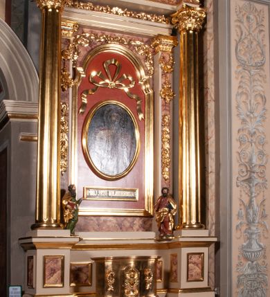 Zdjęcie nr 1: Ołtarz architektoniczny, przyścienny, jednokondygnacyjny, jednoosiowy, w formie podwójnej aediculi. Mensa prostopadłościenna, z płyciną w kształcie leżącego prostokąta wypełnionego marmoryzacją i obwiedzionego profilowaną ramą. Na mensie tabernakulum na planie zbliżonym do owalu, o wklęsło-wypukłej linii ścian. Części boczne na cokole, w środkowej drzwiczki w kształcie stojącego prostokąta zamkniętego łukiem nadwieszonym z symetryczną dekoracją składającą się z kielicha z hostią otoczoną glorią promienistą na tle kłosów zbóż oraz kiści winogron. Na bocznych ściankach dekoracja z ornamentu rocaille'owego. Tabernakulum od góry zamknięte profilowanym gzymsem. 
Nastawa na wysokim, trójdzielnym cokole dekorowanym prostokątnymi płycinami. W polu głównym rama w kształcie stojącego prostokąta zamkniętego od góry łukiem wklęsło-wypukłym z uskokiem. W niej obraz z przedstawieniem Ecce Homo w owalnej ramie poniżej kokardy uwitej ze złotej wstęgi, i powyżej tabliczki z napisem „O MÓJ JEZU MIŁOSIERDZIA!”. Pole flankowane zakończonymi głowicami jońskimi pilastrami podtrzymującymi proste belkowanie. Przed nimi figury dwóch ewangelistów. Całość ujęta dwiema żłobkowanymi kolumnami o kapitelach kompozytowych, wspierającymi pełne belkowanie zwieńczone trójkątnym przyczółkiem z zatkniętym na szczycie krzyżem łacińskim. Na osi kolumn, na belkowaniu wazony z kwiatami. 
Wokół ramy, na trzonach pilastrów, między belkowaniami oraz na fryzie podtrzymywanym przez kolumny dekoracja z elementów roślinnych w postaci pęków kwiatów z liśćmi lub wici; na środku fryzu dwa skrzyżowane młoty; w polu przyczółka uskrzydlona główka anielska. 
Struktura w kolorze kremowym lub marmoryzowana na kolor różowy; kolumny, profile, ornamenty i dekoracje złocone; skrzyżowane młoty, elementy kwiatów i krzyża srebrzone. 