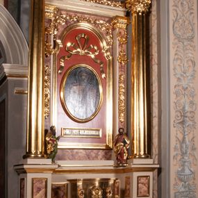 Zdjęcie nr 1: Ołtarz architektoniczny, przyścienny, jednokondygnacyjny, jednoosiowy, w formie podwójnej aediculi. Mensa prostopadłościenna, z płyciną w kształcie leżącego prostokąta wypełnionego marmoryzacją i obwiedzionego profilowaną ramą. Na mensie tabernakulum na planie zbliżonym do owalu, o wklęsło-wypukłej linii ścian. Części boczne na cokole, w środkowej drzwiczki w kształcie stojącego prostokąta zamkniętego łukiem nadwieszonym z symetryczną dekoracją składającą się z kielicha z hostią otoczoną glorią promienistą na tle kłosów zbóż oraz kiści winogron. Na bocznych ściankach dekoracja z ornamentu rocaille&#039;owego. Tabernakulum od góry zamknięte profilowanym gzymsem. 
Nastawa na wysokim, trójdzielnym cokole dekorowanym prostokątnymi płycinami. W polu głównym rama w kształcie stojącego prostokąta zamkniętego od góry łukiem wklęsło-wypukłym z uskokiem. W niej obraz z przedstawieniem Ecce Homo w owalnej ramie poniżej kokardy uwitej ze złotej wstęgi, i powyżej tabliczki z napisem „O MÓJ JEZU MIŁOSIERDZIA!”. Pole flankowane zakończonymi głowicami jońskimi pilastrami podtrzymującymi proste belkowanie. Przed nimi figury dwóch ewangelistów. Całość ujęta dwiema żłobkowanymi kolumnami o kapitelach kompozytowych, wspierającymi pełne belkowanie zwieńczone trójkątnym przyczółkiem z zatkniętym na szczycie krzyżem łacińskim. Na osi kolumn, na belkowaniu wazony z kwiatami. 
Wokół ramy, na trzonach pilastrów, między belkowaniami oraz na fryzie podtrzymywanym przez kolumny dekoracja z elementów roślinnych w postaci pęków kwiatów z liśćmi lub wici; na środku fryzu dwa skrzyżowane młoty; w polu przyczółka uskrzydlona główka anielska. 
Struktura w kolorze kremowym lub marmoryzowana na kolor różowy; kolumny, profile, ornamenty i dekoracje złocone; skrzyżowane młoty, elementy kwiatów i krzyża srebrzone. 