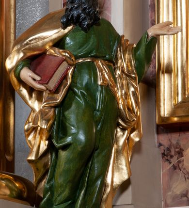 Zdjęcie nr 1: Figura pełna na cokole. Ewangelista ukazany w całej postaci, stojący frontalnie, w kontrapoście, z prawą nogą zgiętą w kolanie i stopą odstawioną nieznacznie do tyłu. Prawą ręką przytrzymuje opartą o biodro zamkniętą księgę, lewa, zgięta w łokciu, uniesiona jest do góry z otwartą, płasko ułożoną dłonią; święty patrzy przed siebie. Ma owalną twarz o okrągłych, szeroko otwartych oczach, ponad którymi widoczne uniesione brwi, o prostym nosie i szerokich ustach; na policzkach rumieńce. Włosy opadają mu na kark, długa broda ułożona w grube pukle rozwiewa się delikatnie na boki. Ewangelista jest ubrany w zieloną suknię przewiązaną w pasie złotym sznurem, zmarszczoną powyżej tego przewiązania; jej fałdy podkreślają ułożenie nóg. Przez plecy i prawą rękę święty ma przerzucony złoty płaszcz. Włosy i zarost brązowe, polichromia w odsłoniętych partiach  karnacji traktowana naturalistycznie.