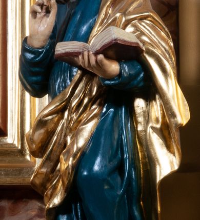 Zdjęcie nr 2: Figura pełna na cokole. Ewangelista ukazany w całej postaci, stojący frontalnie (choć przesunięcie cokołu powoduje, że postać sprawia wrażenie skierowanej w bok), w kontrapoście, z lewą nogą zgiętą w kolanie, jej stopą odsuniętą do tyłu, opartą na palcach. Prawą rękę święty unosi do góry na wysokość piersi, lewą, zgiętą w łokciu podtrzymuje otwartą, ułożoną poziomo księgę; patrzy przed siebie. Ma owalną twarz o okrągłych, szeroko otwartych oczach, ponad którymi widoczne uniesione brwi, o prostym nosie i szerokich ustach; na policzkach rumieńce. Włosy zwinięte w pukle zasłaniają uszy, grube wąsy i jakby rozwiana broda okalają twarz. Ewangelista jest ubrany w niebieską suknię, która grubymi fałdami marszczy się na rękawach, a w części głównej równoległymi fałdami opada w dół, zawijając się nieznacznie w lewą stronę na wysokości kostek. Skośnie przez przód i plecy ma zarzucony złoty płaszcz. Włosy i zarost brązowe, polichromia w odsłoniętych partiach karnacji traktowana naturalistycznie.