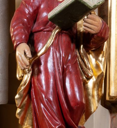 Zdjęcie nr 1: Figura pełna na cokole. Święty Jan ukazany w całej postaci, stojący frontalnie (choć przesunięcie cokołu powoduje, że postać można odbierać jako skierowaną w bok), z prawą nogą wysuniętą do przodu, jakby w trakcie wykonywania kroku w przód lub skrętu. Prawą rękę ma opuszczoną w dół, lewą, zgiętą w łokciu podtrzymuje otwartą księgę; patrzy przed siebie. Święty ma młodzieńczą, owalną twarz z migdałowymi oczami, prostym nosem i małymi ustami, na policzkach widoczne są rumieńce. Długie włosy zwinięte w pukle opadają mu na ramiona i plecy. Ubrany jest w czerwoną suknię, która równoległymi fałdami opada w dół, zawijając się w lewą stronę na wysokości łydek, dzięki czemu podkreśla dynamiczny ruch. Na plecy i lewą rękę ma zarzucony złoty płaszcz spięty złotym sznurem biegnącym na skos przez pierś. Włosy brązowe, polichromia w odsłoniętych partiach ciała karnacji traktowana naturalistycznie.