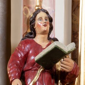 Zdjęcie nr 1: Figura pełna na cokole. Święty Jan ukazany w całej postaci, stojący frontalnie (choć przesunięcie cokołu powoduje, że postać można odbierać jako skierowaną w bok), z prawą nogą wysuniętą do przodu, jakby w trakcie wykonywania kroku w przód lub skrętu. Prawą rękę ma opuszczoną w dół, lewą, zgiętą w łokciu podtrzymuje otwartą księgę; patrzy przed siebie. Święty ma młodzieńczą, owalną twarz z migdałowymi oczami, prostym nosem i małymi ustami, na policzkach widoczne są rumieńce. Długie włosy zwinięte w pukle opadają mu na ramiona i plecy. Ubrany jest w czerwoną suknię, która równoległymi fałdami opada w dół, zawijając się w lewą stronę na wysokości łydek, dzięki czemu podkreśla dynamiczny ruch. Na plecy i lewą rękę ma zarzucony złoty płaszcz spięty złotym sznurem biegnącym na skos przez pierś. Włosy brązowe, polichromia w odsłoniętych partiach ciała karnacji traktowana naturalistycznie.