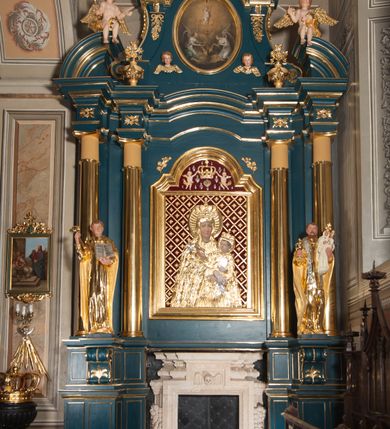 Zdjęcie nr 2: Obraz w kształcie pionowego owalu. W centrum kompozycji Dzieciątko Jezus stojące na monogramie IHS, które lewą ręką przytrzymuje krzyż, a prawą, uniesioną do góry błogosławi. Ma okrągłą twarz i krótkie, jasne włosy, skośnie przez pierś i między nogami biegnie biało-czerwona szarfa, która rozwiewa się za jego plecami. Poniżej monogramu serce Jezusa otoczone koroną cierniową, z płomieniami. Przedstawienie adorowane przez dwa, symetrycznie klęczące anioły z kadzielnicami w rękach. Anioł z prawej strony spogląda w dół, anioł z lewej unosi wzrok w kierunku serca. Oba ubrane są w białe suknie z dużym dekoltem, o szerokich rękawach spiętych złotymi klamrami na wysokości łokci oraz ramion. Każdy ma narzucony niebieski płaszcz i rozpostarte, tonące w mroku skrzydła. Tło wypełniają skłębione chmury w złotobrunatnym odcieniu, które rozjaśniają się za postacią Dzieciątka. 