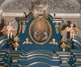 Zdjęcie nr 1: Obraz w kształcie pionowego owalu. W centrum kompozycji Dzieciątko Jezus stojące na monogramie IHS, które lewą ręką przytrzymuje krzyż, a prawą, uniesioną do góry błogosławi. Ma okrągłą twarz i krótkie, jasne włosy, skośnie przez pierś i między nogami biegnie biało-czerwona szarfa, która rozwiewa się za jego plecami. Poniżej monogramu serce Jezusa otoczone koroną cierniową, z płomieniami. Przedstawienie adorowane przez dwa, symetrycznie klęczące anioły z kadzielnicami w rękach. Anioł z prawej strony spogląda w dół, anioł z lewej unosi wzrok w kierunku serca. Oba ubrane są w białe suknie z dużym dekoltem, o szerokich rękawach spiętych złotymi klamrami na wysokości łokci oraz ramion. Każdy ma narzucony niebieski płaszcz i rozpostarte, tonące w mroku skrzydła. Tło wypełniają skłębione chmury w złotobrunatnym odcieniu, które rozjaśniają się za postacią Dzieciątka. 