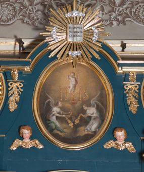 Zdjęcie nr 1: Obraz w kształcie pionowego owalu. W centrum kompozycji Dzieciątko Jezus stojące na monogramie IHS, które lewą ręką przytrzymuje krzyż, a prawą, uniesioną do góry błogosławi. Ma okrągłą twarz i krótkie, jasne włosy, skośnie przez pierś i między nogami biegnie biało-czerwona szarfa, która rozwiewa się za jego plecami. Poniżej monogramu serce Jezusa otoczone koroną cierniową, z płomieniami. Przedstawienie adorowane przez dwa, symetrycznie klęczące anioły z kadzielnicami w rękach. Anioł z prawej strony spogląda w dół, anioł z lewej unosi wzrok w kierunku serca. Oba ubrane są w białe suknie z dużym dekoltem, o szerokich rękawach spiętych złotymi klamrami na wysokości łokci oraz ramion. Każdy ma narzucony niebieski płaszcz i rozpostarte, tonące w mroku skrzydła. Tło wypełniają skłębione chmury w złotobrunatnym odcieniu, które rozjaśniają się za postacią Dzieciątka. 