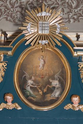 Zdjęcie nr 1: Obraz w kształcie pionowego owalu. W centrum kompozycji Dzieciątko Jezus stojące na monogramie IHS, które lewą ręką przytrzymuje krzyż, a prawą, uniesioną do góry błogosławi. Ma okrągłą twarz i krótkie, jasne włosy, skośnie przez pierś i między nogami biegnie biało-czerwona szarfa, która rozwiewa się za jego plecami. Poniżej monogramu serce Jezusa otoczone koroną cierniową, z płomieniami. Przedstawienie adorowane przez dwa, symetrycznie klęczące anioły z kadzielnicami w rękach. Anioł z prawej strony spogląda w dół, anioł z lewej unosi wzrok w kierunku serca. Oba ubrane są w białe suknie z dużym dekoltem, o szerokich rękawach spiętych złotymi klamrami na wysokości łokci oraz ramion. Każdy ma narzucony niebieski płaszcz i rozpostarte, tonące w mroku skrzydła. Tło wypełniają skłębione chmury w złotobrunatnym odcieniu, które rozjaśniają się za postacią Dzieciątka. 