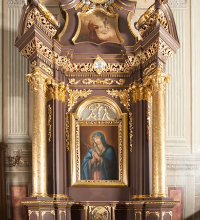 Zdjęcie nr 1: Ołtarz przyścienny, architektoniczny, jednoosiowy, jednokondygnacjowy ze zwieńczeniem. Retabulum ustawione na wysokim cokole, przed którym stoi mensa w typie sarkofagowym. Na mensie tabernakulum w formie prostopadłościanu, którego ściana frontowa jest flankowana parą dostawionych skośnie kolumn, a tylna wolutami. Tabernakulum na cokole, na ścianie frontowej drzwi w kształcie prostokąta zamkniętego od góry łukiem nadwieszonym, z przedstawieniem wypukłorzeźbionej, silnie plastycznej sceny chrztu Chrystusa. Z prawej strony św. Jan Chrzciciel, który prawym kolanem klęczy na głazie, prawą ręką polewa wodą głowę stojącego po lewej stronie Chrystusa. Za nimi dwie postaci, powyżej gołębica Ducha Świętego. W górnych narożnikach ściany frontowej tabernakulum dwa, symetrycznie ułożone ornamenty rocaille. Główne pole właściwego retabulum flankują kolumny korynckie o trzonach dekorowanych wicią, ustawione na tle zwielokrotnionych pilastrów; podpory posadowione na ukośnie ustawionych, rozsuniętych postumentach dekorowanych kwadratowymi płycinami, dźwigają  belkowanie przełamane na ich osiach, z odcinkami także ustawionymi skośnie. W prostokątnym polu głównym nastawy ołtarzowej obraz z przedstawieniem Matki Boskiej Bolesnej poniżej monogramu maryjnego uwitego ze wstęgi, w prostokątnej ramie zamkniętej od góry łukiem nadwieszonym. Górne naroża pola wypełniają płomieniste motywy rocaille przechodzące w dół w fantazyjną wić roślinną.  Na fryzie belkowania umieszczono ornament roślinny z muszlami, a dolna część gzymsu ma formę pasa ażurowej dekoracji z motywów rocaille na czerwonym tle. Na środku tej dekoracji niewielki kartusz z napisem. Na rozsuniętych odcinkach belkowania, na osi kolumn, umieszczone fragmenty pękniętego półkolistego przyczółka; na nich siedzą anioły. Zwieńczenie umieszczone pomiędzy tymi odcinkami w formie ścianki o ogólnym zarysie trapezu o wklęsłych bokach. Wypełnia ją obraz z przedstawieniem św. Antoniego we wklęsło-wypukłej ramie powtarzającej zarys zwieńczenia, które od góry zamknięte jest dwoma wolutami. W miejscu ich styku, na osi, rzeźbione przedstawienie tronującego Boga Ojca pośród chmur, powyżej główek anielskich i na tle glorii promienistej. Bóg prawą rękę wznosi do góry, lewą trzyma na jabłku królewskim i przytrzymuje berło, u stóp gołębica Ducha Świętego. Struktura ołtarza malowana na kolor brązowy; profile, detale, ornamenty i kolumny złocone, postacie w partiach karnacji  polichromowane naturalistycznie.