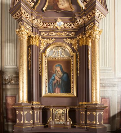 Zdjęcie nr 2: Ołtarz przyścienny, architektoniczny, jednoosiowy, jednokondygnacjowy ze zwieńczeniem. Retabulum ustawione na wysokim cokole, przed którym stoi mensa w typie sarkofagowym. Na mensie tabernakulum w formie prostopadłościanu, którego ściana frontowa jest flankowana parą dostawionych skośnie kolumn, a tylna wolutami. Tabernakulum na cokole, na ścianie frontowej drzwi w kształcie prostokąta zamkniętego od góry łukiem nadwieszonym, z przedstawieniem wypukłorzeźbionej, silnie plastycznej sceny chrztu Chrystusa. Z prawej strony św. Jan Chrzciciel, który prawym kolanem klęczy na głazie, prawą ręką polewa wodą głowę stojącego po lewej stronie Chrystusa. Za nimi dwie postaci, powyżej gołębica Ducha Świętego. W górnych narożnikach ściany frontowej tabernakulum dwa, symetrycznie ułożone ornamenty rocaille. Główne pole właściwego retabulum flankują kolumny korynckie o trzonach dekorowanych wicią, ustawione na tle zwielokrotnionych pilastrów; podpory posadowione na ukośnie ustawionych, rozsuniętych postumentach dekorowanych kwadratowymi płycinami, dźwigają  belkowanie przełamane na ich osiach, z odcinkami także ustawionymi skośnie. W prostokątnym polu głównym nastawy ołtarzowej obraz z przedstawieniem Matki Boskiej Bolesnej poniżej monogramu maryjnego uwitego ze wstęgi, w prostokątnej ramie zamkniętej od góry łukiem nadwieszonym. Górne naroża pola wypełniają płomieniste motywy rocaille przechodzące w dół w fantazyjną wić roślinną.  Na fryzie belkowania umieszczono ornament roślinny z muszlami, a dolna część gzymsu ma formę pasa ażurowej dekoracji z motywów rocaille na czerwonym tle. Na środku tej dekoracji niewielki kartusz z napisem. Na rozsuniętych odcinkach belkowania, na osi kolumn, umieszczone fragmenty pękniętego półkolistego przyczółka; na nich siedzą anioły. Zwieńczenie umieszczone pomiędzy tymi odcinkami w formie ścianki o ogólnym zarysie trapezu o wklęsłych bokach. Wypełnia ją obraz z przedstawieniem św. Antoniego we wklęsło-wypukłej ramie powtarzającej zarys zwieńczenia, które od góry zamknięte jest dwoma wolutami. W miejscu ich styku, na osi, rzeźbione przedstawienie tronującego Boga Ojca pośród chmur, powyżej główek anielskich i na tle glorii promienistej. Bóg prawą rękę wznosi do góry, lewą trzyma na jabłku królewskim i przytrzymuje berło, u stóp gołębica Ducha Świętego. Struktura ołtarza malowana na kolor brązowy; profile, detale, ornamenty i kolumny złocone, postacie w partiach karnacji  polichromowane naturalistycznie.