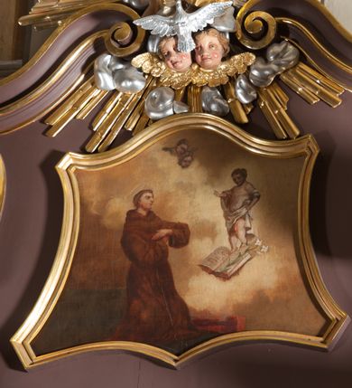 Zdjęcie nr 1: Obraz w kształcie zbliżonym do trapezu o krawędziach bocznych lekko wklęsłych, a dolnej i górnej wklęsło-wypukłej. Obraz przedstawia wizję św. Antoniego. Święty usytuowany nieco na lewo od osi kompozycji, zwrócony w trzech czwartych w lewo, klęczy na ziemi, ręce  założone ma przed sobą na wysokości klatki piersiowej i patrzy na ukazujące mu się Dzieciątko. Antoni ma małą głowę z dużymi oczami, nosem i cofniętym podbródkiem. Krótkie, brązowe włosy przysłaniają ucho, na brodzie krótki zarost. Ubrany w brązowy, obszerny habit z kapturem, łamiący się w grube fałdy. Przed nim, po prawej stronie obrazu u góry Dzieciątko. Chrystus unosi się ponad otwartą księgą i złożonym na niej kwiatem lilii. Prawą nogę wyciąga do przodu, lewą, zgiętą w kolanie ma cofniętą, prawą rękę wyciąga w stronę św. Antoniego w geście błogosławieństwa, lewą podtrzymuje białą tkaninę, która przechodzi przez obie ręce, zasłania przód ciała i regularnymi fałdami opada wzdłuż lewej nogi. Tło wypełniają jasnożółte obłoki na tle ciemniejszego nieba, u góry dwie główki anielskie. Obraz w złoconej, profilowanej ramie.