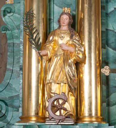 Zdjęcie nr 1: Rzeźba pełna, postać umieszczona na cokole; święta ustawiona w kontrapoście, z lewą nogą wysuniętą do przodu. W prawej dłoni trzyma palmę męczeńską, lewą ugiętą podtrzymuje fałdy płaszcza. Twarz owalna, bez wyrazu, oczy małe, nos prosty, niewielkie usta; falowane, brązowe włosy spływają na plecy, jeden pukiel przechodzi do przodu; na głowie korona. Święta jest ubrana w bogato fałdowaną suknię z gorsetem podkreślającą kształt ciała. Pod szyją ma dwa sznury korali, na jednym zawieszony medalion. Okrywający suknię płaszcz zarzucony na ramiona i plecy opada do ziemi, jedna część przerzucona przez lewą rękę. Spod sukni wystają fragmenty brązowych butów. U dołu, przed świętą połamane koło. 
Szaty i korona złocone, w partiach ciała polichromia naturalistyczna, liść palmy zielony, koło brązowe.