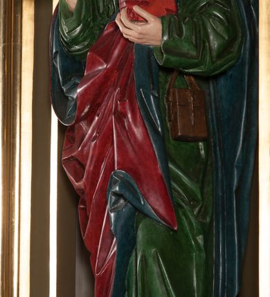 Zdjęcie nr 2: Rzeźba pełna, na cokole. Święty przedstawiony w całej postaci, w pozycji stojącej w ujęciu frontalnym, w lekkim kontrapoście, z lewą nogą wysuniętą do przodu. Ma uniesione ręce – prawą przed siebie, z dłonią otwartą, w lewej trzyma zamkniętą księgę. Głowę ma uniesioną ku górze, nieznacznie przechyloną na lewo. Ma młodzieńczą twarz o wyrazistych rysach: szerokie, delikatnie przymknięte oczy pod uniesionymi, wąskimi brwiami, długi nos, szerokie usta o kącikach skierowanych w dół oraz silnie wysunięte kości policzkowe. Długie włosy, ciasno zwinięte w pukle, opadają na czoło i kark. Święty jest ubrany w przepasaną, rozciętą pod szyją zieloną suknię oraz spięty pod szyją czerwony płaszcz z niebieskim podbiciem. Szaty układają się w głębokie, ostro łamiące się fałdy. Płaszcz ponad lewym kolanem odwija się w formę rozetki; na lewym boku przytroczona do pasa, prostokątna, brązowa sakiewka. Polichromia w partiach ciała naturalistyczna. 