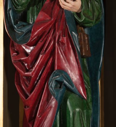 Zdjęcie nr 1: Rzeźba pełna, na cokole. Święty przedstawiony w całej postaci, w pozycji stojącej w ujęciu frontalnym, w lekkim kontrapoście, z lewą nogą wysuniętą do przodu. Ma uniesione ręce – prawą przed siebie, z dłonią otwartą, w lewej trzyma zamkniętą księgę. Głowę ma uniesioną ku górze, nieznacznie przechyloną na lewo. Ma młodzieńczą twarz o wyrazistych rysach: szerokie, delikatnie przymknięte oczy pod uniesionymi, wąskimi brwiami, długi nos, szerokie usta o kącikach skierowanych w dół oraz silnie wysunięte kości policzkowe. Długie włosy, ciasno zwinięte w pukle, opadają na czoło i kark. Święty jest ubrany w przepasaną, rozciętą pod szyją zieloną suknię oraz spięty pod szyją czerwony płaszcz z niebieskim podbiciem. Szaty układają się w głębokie, ostro łamiące się fałdy. Płaszcz ponad lewym kolanem odwija się w formę rozetki; na lewym boku przytroczona do pasa, prostokątna, brązowa sakiewka. Polichromia w partiach ciała naturalistyczna. 