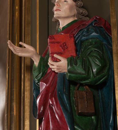 Zdjęcie nr 4: Rzeźba pełna, na cokole. Święty przedstawiony w całej postaci, w pozycji stojącej w ujęciu frontalnym, w lekkim kontrapoście, z lewą nogą wysuniętą do przodu. Ma uniesione ręce – prawą przed siebie, z dłonią otwartą, w lewej trzyma zamkniętą księgę. Głowę ma uniesioną ku górze, nieznacznie przechyloną na lewo. Ma młodzieńczą twarz o wyrazistych rysach: szerokie, delikatnie przymknięte oczy pod uniesionymi, wąskimi brwiami, długi nos, szerokie usta o kącikach skierowanych w dół oraz silnie wysunięte kości policzkowe. Długie włosy, ciasno zwinięte w pukle, opadają na czoło i kark. Święty jest ubrany w przepasaną, rozciętą pod szyją zieloną suknię oraz spięty pod szyją czerwony płaszcz z niebieskim podbiciem. Szaty układają się w głębokie, ostro łamiące się fałdy. Płaszcz ponad lewym kolanem odwija się w formę rozetki; na lewym boku przytroczona do pasa, prostokątna, brązowa sakiewka. Polichromia w partiach ciała naturalistyczna. 