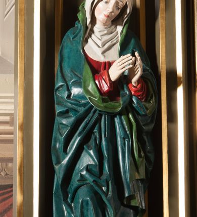 Zdjęcie nr 2: Rzeźba pełna, na cokole. Maria przedstawiona w całej postaci, w pozycji stojącej w ujęciu frontalnym. Ręce składa do modlitwy, głowę ma pochyloną na lewe ramię, spogląda w dół. Na jej łagodnej twarzy maluje się smutek, spod uniesionych brwi spoglądają duże, szare oczy o zewnętrznych kącikach skierowany w dół, nos mały, usta drobne, czerwone; na policzkach rumieńce. 
Maria jest ubrana w długą, czerwoną suknię. Głowę okrywa  biały welon, a dekolt tego samego koloru podwika. Na suknię ma narzucony niebieski płaszcz z zielonym podbiciem, który układa się w gęste, głębokie fałdy i wywija w formę kołnierza, wokół dłoni oraz tworzy małą rozetkę przy prawej stopie. Spod sukni wystają szpiczaste pantofelki. Polichromia w partiach ciała naturalistyczna. 
