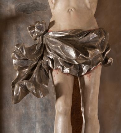 Zdjęcie nr 6: Rzeźba pełna, całopostaciowa. Chrystus rozpięty na krzyżu, ciało wyprężone, z podkreśloną linią żeber i słabo zaznaczoną muskulaturą. Głowa w cierniowej koronie opada silnie na prawe ramię; usta rozchylone, oczy przymknięte. Twarz podłużna, okolona krótkim zarostem; długie, falowane włosy z zaznaczonymi kosmykami opadają na kark i plecy, jeden pukiel do przodu na klatkę piersiową. Ręce rozciągnięte skośnie do góry, przybite do ramion krzyża gwoździami, palce dłoni podwinięte. Klatka piersiowa wysunięta ku przodowi, o wyraźnie zaznaczonej anatomii. Nogi proste, ułożone sztywno; stopy skrzyżowane i przebite jednym gwoździem. Na ciele słabo widoczne ślady męki – rana w boku, krew wypływająca z miejsc przebicia gwoździami oraz spod korony cierniowej. Krótkie perizonium zakrywa biodra, jest związane na prawym boku i rozwiewa się przy prawym udzie. Jest obficie drapowane w głębokie, drobne fałdy. Chrystus jest przybity do krzyża bejcowanego na ciemny kolor, otoczonego złotą ramą. Na jego górnej belce titulus z inskrypcją „INRI” w formie przybitego gwoździem, rozwijającego się zwoju z rozciętą dolną krawędzią. W tle, do wysokości kostek Chrystusa wieloplanowa, płaskorzeźbiona panorama Jerozolimy, powyżej niebo z plastycznie zaznaczonymi obłokami. 
Polichromia w partiach ciała naturalistyczna, perizonium i tło srebrzone. 
