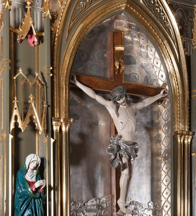 Zdjęcie nr 3: Rzeźba pełna, całopostaciowa. Chrystus rozpięty na krzyżu, ciało wyprężone, z podkreśloną linią żeber i słabo zaznaczoną muskulaturą. Głowa w cierniowej koronie opada silnie na prawe ramię; usta rozchylone, oczy przymknięte. Twarz podłużna, okolona krótkim zarostem; długie, falowane włosy z zaznaczonymi kosmykami opadają na kark i plecy, jeden pukiel do przodu na klatkę piersiową. Ręce rozciągnięte skośnie do góry, przybite do ramion krzyża gwoździami, palce dłoni podwinięte. Klatka piersiowa wysunięta ku przodowi, o wyraźnie zaznaczonej anatomii. Nogi proste, ułożone sztywno; stopy skrzyżowane i przebite jednym gwoździem. Na ciele słabo widoczne ślady męki – rana w boku, krew wypływająca z miejsc przebicia gwoździami oraz spod korony cierniowej. Krótkie perizonium zakrywa biodra, jest związane na prawym boku i rozwiewa się przy prawym udzie. Jest obficie drapowane w głębokie, drobne fałdy. Chrystus jest przybity do krzyża bejcowanego na ciemny kolor, otoczonego złotą ramą. Na jego górnej belce titulus z inskrypcją „INRI” w formie przybitego gwoździem, rozwijającego się zwoju z rozciętą dolną krawędzią. W tle, do wysokości kostek Chrystusa wieloplanowa, płaskorzeźbiona panorama Jerozolimy, powyżej niebo z plastycznie zaznaczonymi obłokami. 
Polichromia w partiach ciała naturalistyczna, perizonium i tło srebrzone. 
