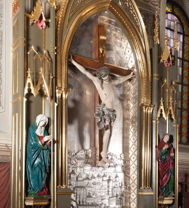 Zdjęcie nr 9: Rzeźba pełna, całopostaciowa. Chrystus rozpięty na krzyżu, ciało wyprężone, z podkreśloną linią żeber i słabo zaznaczoną muskulaturą. Głowa w cierniowej koronie opada silnie na prawe ramię; usta rozchylone, oczy przymknięte. Twarz podłużna, okolona krótkim zarostem; długie, falowane włosy z zaznaczonymi kosmykami opadają na kark i plecy, jeden pukiel do przodu na klatkę piersiową. Ręce rozciągnięte skośnie do góry, przybite do ramion krzyża gwoździami, palce dłoni podwinięte. Klatka piersiowa wysunięta ku przodowi, o wyraźnie zaznaczonej anatomii. Nogi proste, ułożone sztywno; stopy skrzyżowane i przebite jednym gwoździem. Na ciele słabo widoczne ślady męki – rana w boku, krew wypływająca z miejsc przebicia gwoździami oraz spod korony cierniowej. Krótkie perizonium zakrywa biodra, jest związane na prawym boku i rozwiewa się przy prawym udzie. Jest obficie drapowane w głębokie, drobne fałdy. Chrystus jest przybity do krzyża bejcowanego na ciemny kolor, otoczonego złotą ramą. Na jego górnej belce titulus z inskrypcją „INRI” w formie przybitego gwoździem, rozwijającego się zwoju z rozciętą dolną krawędzią. W tle, do wysokości kostek Chrystusa wieloplanowa, płaskorzeźbiona panorama Jerozolimy, powyżej niebo z plastycznie zaznaczonymi obłokami. 
Polichromia w partiach ciała naturalistyczna, perizonium i tło srebrzone. 
