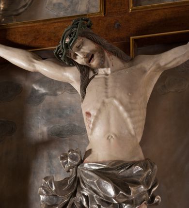 Zdjęcie nr 1: Rzeźba pełna, całopostaciowa. Chrystus rozpięty na krzyżu, ciało wyprężone, z podkreśloną linią żeber i słabo zaznaczoną muskulaturą. Głowa w cierniowej koronie opada silnie na prawe ramię; usta rozchylone, oczy przymknięte. Twarz podłużna, okolona krótkim zarostem; długie, falowane włosy z zaznaczonymi kosmykami opadają na kark i plecy, jeden pukiel do przodu na klatkę piersiową. Ręce rozciągnięte skośnie do góry, przybite do ramion krzyża gwoździami, palce dłoni podwinięte. Klatka piersiowa wysunięta ku przodowi, o wyraźnie zaznaczonej anatomii. Nogi proste, ułożone sztywno; stopy skrzyżowane i przebite jednym gwoździem. Na ciele słabo widoczne ślady męki – rana w boku, krew wypływająca z miejsc przebicia gwoździami oraz spod korony cierniowej. Krótkie perizonium zakrywa biodra, jest związane na prawym boku i rozwiewa się przy prawym udzie. Jest obficie drapowane w głębokie, drobne fałdy. Chrystus jest przybity do krzyża bejcowanego na ciemny kolor, otoczonego złotą ramą. Na jego górnej belce titulus z inskrypcją „INRI” w formie przybitego gwoździem, rozwijającego się zwoju z rozciętą dolną krawędzią. W tle, do wysokości kostek Chrystusa wieloplanowa, płaskorzeźbiona panorama Jerozolimy, powyżej niebo z plastycznie zaznaczonymi obłokami. 
Polichromia w partiach ciała naturalistyczna, perizonium i tło srebrzone. 

