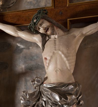 Zdjęcie nr 1: Rzeźba pełna, całopostaciowa. Chrystus rozpięty na krzyżu, ciało wyprężone, z podkreśloną linią żeber i słabo zaznaczoną muskulaturą. Głowa w cierniowej koronie opada silnie na prawe ramię; usta rozchylone, oczy przymknięte. Twarz podłużna, okolona krótkim zarostem; długie, falowane włosy z zaznaczonymi kosmykami opadają na kark i plecy, jeden pukiel do przodu na klatkę piersiową. Ręce rozciągnięte skośnie do góry, przybite do ramion krzyża gwoździami, palce dłoni podwinięte. Klatka piersiowa wysunięta ku przodowi, o wyraźnie zaznaczonej anatomii. Nogi proste, ułożone sztywno; stopy skrzyżowane i przebite jednym gwoździem. Na ciele słabo widoczne ślady męki – rana w boku, krew wypływająca z miejsc przebicia gwoździami oraz spod korony cierniowej. Krótkie perizonium zakrywa biodra, jest związane na prawym boku i rozwiewa się przy prawym udzie. Jest obficie drapowane w głębokie, drobne fałdy. Chrystus jest przybity do krzyża bejcowanego na ciemny kolor, otoczonego złotą ramą. Na jego górnej belce titulus z inskrypcją „INRI” w formie przybitego gwoździem, rozwijającego się zwoju z rozciętą dolną krawędzią. W tle, do wysokości kostek Chrystusa wieloplanowa, płaskorzeźbiona panorama Jerozolimy, powyżej niebo z plastycznie zaznaczonymi obłokami. 
Polichromia w partiach ciała naturalistyczna, perizonium i tło srebrzone. 
