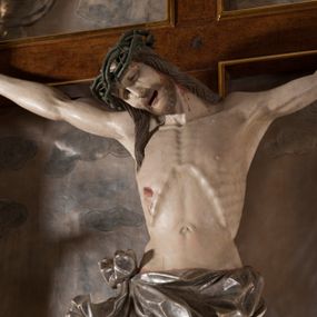 Zdjęcie nr 1: Rzeźba pełna, całopostaciowa. Chrystus rozpięty na krzyżu, ciało wyprężone, z podkreśloną linią żeber i słabo zaznaczoną muskulaturą. Głowa w cierniowej koronie opada silnie na prawe ramię; usta rozchylone, oczy przymknięte. Twarz podłużna, okolona krótkim zarostem; długie, falowane włosy z zaznaczonymi kosmykami opadają na kark i plecy, jeden pukiel do przodu na klatkę piersiową. Ręce rozciągnięte skośnie do góry, przybite do ramion krzyża gwoździami, palce dłoni podwinięte. Klatka piersiowa wysunięta ku przodowi, o wyraźnie zaznaczonej anatomii. Nogi proste, ułożone sztywno; stopy skrzyżowane i przebite jednym gwoździem. Na ciele słabo widoczne ślady męki – rana w boku, krew wypływająca z miejsc przebicia gwoździami oraz spod korony cierniowej. Krótkie perizonium zakrywa biodra, jest związane na prawym boku i rozwiewa się przy prawym udzie. Jest obficie drapowane w głębokie, drobne fałdy. Chrystus jest przybity do krzyża bejcowanego na ciemny kolor, otoczonego złotą ramą. Na jego górnej belce titulus z inskrypcją „INRI” w formie przybitego gwoździem, rozwijającego się zwoju z rozciętą dolną krawędzią. W tle, do wysokości kostek Chrystusa wieloplanowa, płaskorzeźbiona panorama Jerozolimy, powyżej niebo z plastycznie zaznaczonymi obłokami. 
Polichromia w partiach ciała naturalistyczna, perizonium i tło srebrzone. 
