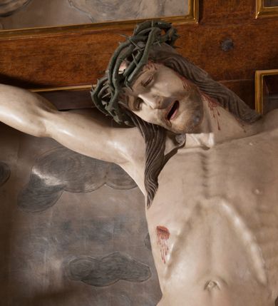 Zdjęcie nr 4: Rzeźba pełna, całopostaciowa. Chrystus rozpięty na krzyżu, ciało wyprężone, z podkreśloną linią żeber i słabo zaznaczoną muskulaturą. Głowa w cierniowej koronie opada silnie na prawe ramię; usta rozchylone, oczy przymknięte. Twarz podłużna, okolona krótkim zarostem; długie, falowane włosy z zaznaczonymi kosmykami opadają na kark i plecy, jeden pukiel do przodu na klatkę piersiową. Ręce rozciągnięte skośnie do góry, przybite do ramion krzyża gwoździami, palce dłoni podwinięte. Klatka piersiowa wysunięta ku przodowi, o wyraźnie zaznaczonej anatomii. Nogi proste, ułożone sztywno; stopy skrzyżowane i przebite jednym gwoździem. Na ciele słabo widoczne ślady męki – rana w boku, krew wypływająca z miejsc przebicia gwoździami oraz spod korony cierniowej. Krótkie perizonium zakrywa biodra, jest związane na prawym boku i rozwiewa się przy prawym udzie. Jest obficie drapowane w głębokie, drobne fałdy. Chrystus jest przybity do krzyża bejcowanego na ciemny kolor, otoczonego złotą ramą. Na jego górnej belce titulus z inskrypcją „INRI” w formie przybitego gwoździem, rozwijającego się zwoju z rozciętą dolną krawędzią. W tle, do wysokości kostek Chrystusa wieloplanowa, płaskorzeźbiona panorama Jerozolimy, powyżej niebo z plastycznie zaznaczonymi obłokami. 
Polichromia w partiach ciała naturalistyczna, perizonium i tło srebrzone. 
