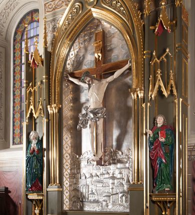 Zdjęcie nr 2: Rzeźba pełna, całopostaciowa. Chrystus rozpięty na krzyżu, ciało wyprężone, z podkreśloną linią żeber i słabo zaznaczoną muskulaturą. Głowa w cierniowej koronie opada silnie na prawe ramię; usta rozchylone, oczy przymknięte. Twarz podłużna, okolona krótkim zarostem; długie, falowane włosy z zaznaczonymi kosmykami opadają na kark i plecy, jeden pukiel do przodu na klatkę piersiową. Ręce rozciągnięte skośnie do góry, przybite do ramion krzyża gwoździami, palce dłoni podwinięte. Klatka piersiowa wysunięta ku przodowi, o wyraźnie zaznaczonej anatomii. Nogi proste, ułożone sztywno; stopy skrzyżowane i przebite jednym gwoździem. Na ciele słabo widoczne ślady męki – rana w boku, krew wypływająca z miejsc przebicia gwoździami oraz spod korony cierniowej. Krótkie perizonium zakrywa biodra, jest związane na prawym boku i rozwiewa się przy prawym udzie. Jest obficie drapowane w głębokie, drobne fałdy. Chrystus jest przybity do krzyża bejcowanego na ciemny kolor, otoczonego złotą ramą. Na jego górnej belce titulus z inskrypcją „INRI” w formie przybitego gwoździem, rozwijającego się zwoju z rozciętą dolną krawędzią. W tle, do wysokości kostek Chrystusa wieloplanowa, płaskorzeźbiona panorama Jerozolimy, powyżej niebo z plastycznie zaznaczonymi obłokami. 
Polichromia w partiach ciała naturalistyczna, perizonium i tło srebrzone. 
