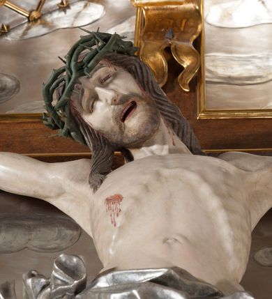 Zdjęcie nr 5: Rzeźba pełna, całopostaciowa. Chrystus rozpięty na krzyżu, ciało wyprężone, z podkreśloną linią żeber i słabo zaznaczoną muskulaturą. Głowa w cierniowej koronie opada silnie na prawe ramię; usta rozchylone, oczy przymknięte. Twarz podłużna, okolona krótkim zarostem; długie, falowane włosy z zaznaczonymi kosmykami opadają na kark i plecy, jeden pukiel do przodu na klatkę piersiową. Ręce rozciągnięte skośnie do góry, przybite do ramion krzyża gwoździami, palce dłoni podwinięte. Klatka piersiowa wysunięta ku przodowi, o wyraźnie zaznaczonej anatomii. Nogi proste, ułożone sztywno; stopy skrzyżowane i przebite jednym gwoździem. Na ciele słabo widoczne ślady męki – rana w boku, krew wypływająca z miejsc przebicia gwoździami oraz spod korony cierniowej. Krótkie perizonium zakrywa biodra, jest związane na prawym boku i rozwiewa się przy prawym udzie. Jest obficie drapowane w głębokie, drobne fałdy. Chrystus jest przybity do krzyża bejcowanego na ciemny kolor, otoczonego złotą ramą. Na jego górnej belce titulus z inskrypcją „INRI” w formie przybitego gwoździem, rozwijającego się zwoju z rozciętą dolną krawędzią. W tle, do wysokości kostek Chrystusa wieloplanowa, płaskorzeźbiona panorama Jerozolimy, powyżej niebo z plastycznie zaznaczonymi obłokami. 
Polichromia w partiach ciała naturalistyczna, perizonium i tło srebrzone. 
