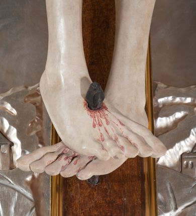 Zdjęcie nr 8: Rzeźba pełna, całopostaciowa. Chrystus rozpięty na krzyżu, ciało wyprężone, z podkreśloną linią żeber i słabo zaznaczoną muskulaturą. Głowa w cierniowej koronie opada silnie na prawe ramię; usta rozchylone, oczy przymknięte. Twarz podłużna, okolona krótkim zarostem; długie, falowane włosy z zaznaczonymi kosmykami opadają na kark i plecy, jeden pukiel do przodu na klatkę piersiową. Ręce rozciągnięte skośnie do góry, przybite do ramion krzyża gwoździami, palce dłoni podwinięte. Klatka piersiowa wysunięta ku przodowi, o wyraźnie zaznaczonej anatomii. Nogi proste, ułożone sztywno; stopy skrzyżowane i przebite jednym gwoździem. Na ciele słabo widoczne ślady męki – rana w boku, krew wypływająca z miejsc przebicia gwoździami oraz spod korony cierniowej. Krótkie perizonium zakrywa biodra, jest związane na prawym boku i rozwiewa się przy prawym udzie. Jest obficie drapowane w głębokie, drobne fałdy. Chrystus jest przybity do krzyża bejcowanego na ciemny kolor, otoczonego złotą ramą. Na jego górnej belce titulus z inskrypcją „INRI” w formie przybitego gwoździem, rozwijającego się zwoju z rozciętą dolną krawędzią. W tle, do wysokości kostek Chrystusa wieloplanowa, płaskorzeźbiona panorama Jerozolimy, powyżej niebo z plastycznie zaznaczonymi obłokami. 
Polichromia w partiach ciała naturalistyczna, perizonium i tło srebrzone. 
