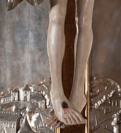 Zdjęcie nr 7: Rzeźba pełna, całopostaciowa. Chrystus rozpięty na krzyżu, ciało wyprężone, z podkreśloną linią żeber i słabo zaznaczoną muskulaturą. Głowa w cierniowej koronie opada silnie na prawe ramię; usta rozchylone, oczy przymknięte. Twarz podłużna, okolona krótkim zarostem; długie, falowane włosy z zaznaczonymi kosmykami opadają na kark i plecy, jeden pukiel do przodu na klatkę piersiową. Ręce rozciągnięte skośnie do góry, przybite do ramion krzyża gwoździami, palce dłoni podwinięte. Klatka piersiowa wysunięta ku przodowi, o wyraźnie zaznaczonej anatomii. Nogi proste, ułożone sztywno; stopy skrzyżowane i przebite jednym gwoździem. Na ciele słabo widoczne ślady męki – rana w boku, krew wypływająca z miejsc przebicia gwoździami oraz spod korony cierniowej. Krótkie perizonium zakrywa biodra, jest związane na prawym boku i rozwiewa się przy prawym udzie. Jest obficie drapowane w głębokie, drobne fałdy. Chrystus jest przybity do krzyża bejcowanego na ciemny kolor, otoczonego złotą ramą. Na jego górnej belce titulus z inskrypcją „INRI” w formie przybitego gwoździem, rozwijającego się zwoju z rozciętą dolną krawędzią. W tle, do wysokości kostek Chrystusa wieloplanowa, płaskorzeźbiona panorama Jerozolimy, powyżej niebo z plastycznie zaznaczonymi obłokami. 
Polichromia w partiach ciała naturalistyczna, perizonium i tło srebrzone. 
