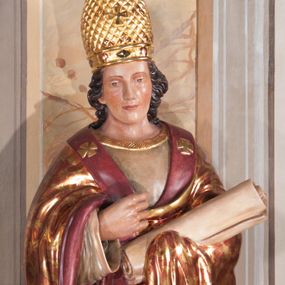 Zdjęcie nr 1: Całopostaciowe przedstawienie świętego Klemensa na cokole. Święty patrzy przed siebie, ma głowę przechyloną lekko na prawą stronę, prawą rękę unosi na wysokość dekoltu, w lewej, osłoniętej płaszczem trzyma zwój. Ma owalną twarz o łagodnym wyrazie, ciemne oczy, szeroki nos i niewielkie, czerwone usta, na policzkach delikatny rumieniec. Jest ubrany w albę z pasem koronki u dołu i obszerną, czerwoną kapę dekorowaną złotymi liśćmi; na głowie ma tiarę, a na nogach buty ze złotymi gwiazdami. 