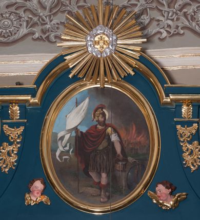 Zdjęcie nr 1: Obraz w kształcie stojącego owalu z przedstawieniem św. Floriana. W centrum kompozycji święty przedstawiony w całej postaci, stojący, zwrócony w trzech czwartych w prawo, w kontrapoście, z lewą noga wysuniętą do przodu. W prawej ręce trzyma białą chorągiew, w lewej cebrzyk, z którego wylewa się woda. Ma owalną twarz o poważnym wyrazie, okoloną gęstym, ciemnym zarostem; wokół głowy żółty nimb. Święty ubrany jest w strój rzymskiego legionisty – złotą zbroję z naramiennikami, nałożoną na białą koszulę, brązową spódnicę i hełm z czerwonym pióropuszem. Na zbroję narzucony ma płaszcz spięty na lewym barku i przełożony przez prawą rękę, układający się w równoległe fałdy. Święty stoi na ziemi, obok niego oparta tarcza. W tle, z prawej strony kompozycji płonie budynek, ponad którym wznoszą się kłęby brunatnego dymu; z lewej strony niebieskie niebo. Obraz w prostej, profilowanej, złoconej ramie.