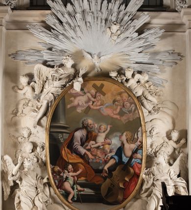 Zdjęcie nr 16: Owalny obraz, w drewnianej, profilowanej, złoconej ramie ukazuje klęczącego św. Józefa z siedzącym na chmurach Jezusem w towarzystwie grającego na kontrabasie anioła, grup uskrzydlonych puttów i główek anielskich. Józef ukazany jest w trzech czwartych, z lewą ręką obejmującą Jezusa, a prawą, zgiętą w łokciu i wyciągniętą do przodu. Głowa z łysiną czołową i siwymi, kręconymi, krótkimi włosami; twarz o rysach starszego mężczyzny, z krótkim nosem i otwartymi oczami. Święty ubrany jest w jasnofioletową suknię i brązowy płaszcz okrywający niemal cała sylwetkę. Siedzący na chmurze z trzema główkami aniołków nagi Jezus głowę zwraca ku górze i wysoko unosi rączki. Jego twarz jest pełna, policzki pękate, zaczerwienione, oczy ciemnobrązowe, krótki nosek i otwarte, pełne usta, włosy krótkie, jasne, kręcone.  Po prawej stronie klęczy anioł grający na kontrabasie, ukazany w trzech czwartych, zwrócony głową w prawo, ubrany w niebieską suknię odsłaniającą lewą pierś i bladoczerwony płaszcz. Jego głowa jest przedstawiona z profilu, z gęstymi, kręconymi, blond włosami, opadającymi na ramiona. Twarz ma pociągłą, z długim, zadartym nosem, otwartymi oczami i różowymi, otwartymi ustami. Przed św. Józefem siedzi para aniołków, z których jeden trzyma długi kij zakończony lilią. W górnej strefie wyłaniają się z chmur główki aniołków i dwa uskrzydlone aniołki trzymające krzyż. Tło nieokreślone, w tonacjach szarości, z widoczną dolną częścią kolumny. 
