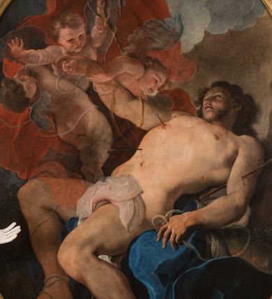 Zdjęcie nr 1: Obraz w kształcie owalu, w profilowanej, złoconej ramie ukazuje na wpół leżącego na skale, niemal nagiego św. Sebastiana przebitego strzałami w towarzystwie uskrzydlonych aniołków. Święty ukazany w trzech czwartych, z silnie wysklepioną klatką piersiową, głowę zwraca ku górze. Jego prawą, uniesioną rękę podtrzymuje anioł, lewa ręka spoczywa na skale. Biodra zasłania biała, zakrwawiona chusta, a na skale rozpostarta jest niebieska szata. Na nogach i rękach widoczne resztki krępujących go sznurów. Ciało przebijają trzy strzały, dwie w klatce piersiowej, jedna w lewym ramieniu. Za świętym trzy nagie niemalże aniołki w zróżnicowanych pozach, z których jeden trzyma palmę męczeństwa; anioły ukazane szkicowo, na tle czerwonej, rozłożystej tkaniny. W tle skłębione niebieskie chmury i szaro-brązowa nieokreślona przestrzeń. Po lewej, u dołu sygnatura „Paolo Pagani Pinxit&quot;.