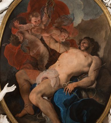 Zdjęcie nr 11: Obraz w kształcie owalu, w profilowanej, złoconej ramie ukazuje na wpół leżącego na skale, niemal nagiego św. Sebastiana przebitego strzałami w towarzystwie uskrzydlonych aniołków. Święty ukazany w trzech czwartych, z silnie wysklepioną klatką piersiową, głowę zwraca ku górze. Jego prawą, uniesioną rękę podtrzymuje anioł, lewa ręka spoczywa na skale. Biodra zasłania biała, zakrwawiona chusta, a na skale rozpostarta jest niebieska szata. Na nogach i rękach widoczne resztki krępujących go sznurów. Ciało przebijają trzy strzały, dwie w klatce piersiowej, jedna w lewym ramieniu. Za świętym trzy nagie niemalże aniołki w zróżnicowanych pozach, z których jeden trzyma palmę męczeństwa; anioły ukazane szkicowo, na tle czerwonej, rozłożystej tkaniny. W tle skłębione niebieskie chmury i szaro-brązowa nieokreślona przestrzeń. Po lewej, u dołu sygnatura „Paolo Pagani Pinxit&quot;.