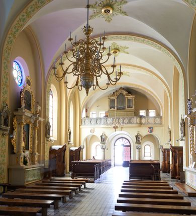 Zdjęcie nr 60: Kościół murowany, orientowany, jednonawowy, trójprzęsłowy, z transeptem i jednoprzęsłowym, węższym i niższym prezbiterium zamkniętym trójbocznie. Między nawą a transeptem po obu stronach znajdują się kruchty, do prezbiterium od strony północnej dostawiona kruchta złożona z dwóch lokalności. Ściany są rozczłonkowane filarami, tworzącymi płytkie arkadowe wnęki. Między nawą a prezbiterium arkada tęczowa. Sklepienie krzyżowe na gurtach spływających na filary. Prezbiterium oświetlają wąskie, prostokątne okna zamknięte ostrołukowo, w nawie i w transepcie znajdują się prostokątne okna prostokątne zamknięte półkoliście, w ścianie frontowej w dolnej części okna prostokątne zamknięte półkoliście, a w górnej podwójne okna prostokątne. Fasada ujęta dwiema narożnymi szkarpami, zwieńczona trójkątnie. Do niej dostawiona kruchta z dwuspadowym daszkiem i wejściem w kształcie prostokąta zamkniętego półkoliście. W kamiennej fasadzie znajdują się dołem okna prostokątne, zamknięte półkoliście, a górą  prostokątne, biforialne okna z nadprożem łukowatym wykonanym z cegły. Szczyt ceglany, podzielony na trzy strefy kamiennym wątkiem, z pięcioma wnękami prostokątnymi, zamkniętymi półkoliście, z wąskimi otworami w dolnej strefie i z owalnym oknem w drugiej. Elewacje boczne nawy ujęte skarpami. Dachy dwuspadowe, kryte dachówką, z wieżyczką na sygnaturkę.         