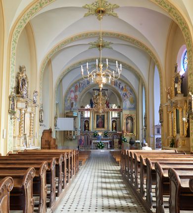 Zdjęcie nr 56: Kościół murowany, orientowany, jednonawowy, trójprzęsłowy, z transeptem i jednoprzęsłowym, węższym i niższym prezbiterium zamkniętym trójbocznie. Między nawą a transeptem po obu stronach znajdują się kruchty, do prezbiterium od strony północnej dostawiona kruchta złożona z dwóch lokalności. Ściany są rozczłonkowane filarami, tworzącymi płytkie arkadowe wnęki. Między nawą a prezbiterium arkada tęczowa. Sklepienie krzyżowe na gurtach spływających na filary. Prezbiterium oświetlają wąskie, prostokątne okna zamknięte ostrołukowo, w nawie i w transepcie znajdują się prostokątne okna prostokątne zamknięte półkoliście, w ścianie frontowej w dolnej części okna prostokątne zamknięte półkoliście, a w górnej podwójne okna prostokątne. Fasada ujęta dwiema narożnymi szkarpami, zwieńczona trójkątnie. Do niej dostawiona kruchta z dwuspadowym daszkiem i wejściem w kształcie prostokąta zamkniętego półkoliście. W kamiennej fasadzie znajdują się dołem okna prostokątne, zamknięte półkoliście, a górą  prostokątne, biforialne okna z nadprożem łukowatym wykonanym z cegły. Szczyt ceglany, podzielony na trzy strefy kamiennym wątkiem, z pięcioma wnękami prostokątnymi, zamkniętymi półkoliście, z wąskimi otworami w dolnej strefie i z owalnym oknem w drugiej. Elewacje boczne nawy ujęte skarpami. Dachy dwuspadowe, kryte dachówką, z wieżyczką na sygnaturkę.         