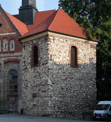 Zdjęcie nr 4: Kościół murowany, orientowany, jednonawowy, trójprzęsłowy, z transeptem i jednoprzęsłowym, węższym i niższym prezbiterium zamkniętym trójbocznie. Między nawą a transeptem po obu stronach znajdują się kruchty, do prezbiterium od strony północnej dostawiona kruchta złożona z dwóch lokalności. Ściany są rozczłonkowane filarami, tworzącymi płytkie arkadowe wnęki. Między nawą a prezbiterium arkada tęczowa. Sklepienie krzyżowe na gurtach spływających na filary. Prezbiterium oświetlają wąskie, prostokątne okna zamknięte ostrołukowo, w nawie i w transepcie znajdują się prostokątne okna prostokątne zamknięte półkoliście, w ścianie frontowej w dolnej części okna prostokątne zamknięte półkoliście, a w górnej podwójne okna prostokątne. Fasada ujęta dwiema narożnymi szkarpami, zwieńczona trójkątnie. Do niej dostawiona kruchta z dwuspadowym daszkiem i wejściem w kształcie prostokąta zamkniętego półkoliście. W kamiennej fasadzie znajdują się dołem okna prostokątne, zamknięte półkoliście, a górą  prostokątne, biforialne okna z nadprożem łukowatym wykonanym z cegły. Szczyt ceglany, podzielony na trzy strefy kamiennym wątkiem, z pięcioma wnękami prostokątnymi, zamkniętymi półkoliście, z wąskimi otworami w dolnej strefie i z owalnym oknem w drugiej. Elewacje boczne nawy ujęte skarpami. Dachy dwuspadowe, kryte dachówką, z wieżyczką na sygnaturkę.         