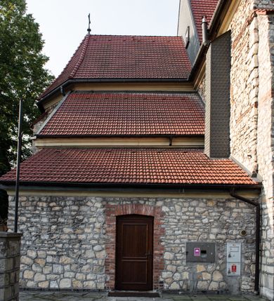 Zdjęcie nr 6: Kościół murowany, orientowany, jednonawowy, trójprzęsłowy, z transeptem i jednoprzęsłowym, węższym i niższym prezbiterium zamkniętym trójbocznie. Między nawą a transeptem po obu stronach znajdują się kruchty, do prezbiterium od strony północnej dostawiona kruchta złożona z dwóch lokalności. Ściany są rozczłonkowane filarami, tworzącymi płytkie arkadowe wnęki. Między nawą a prezbiterium arkada tęczowa. Sklepienie krzyżowe na gurtach spływających na filary. Prezbiterium oświetlają wąskie, prostokątne okna zamknięte ostrołukowo, w nawie i w transepcie znajdują się prostokątne okna prostokątne zamknięte półkoliście, w ścianie frontowej w dolnej części okna prostokątne zamknięte półkoliście, a w górnej podwójne okna prostokątne. Fasada ujęta dwiema narożnymi szkarpami, zwieńczona trójkątnie. Do niej dostawiona kruchta z dwuspadowym daszkiem i wejściem w kształcie prostokąta zamkniętego półkoliście. W kamiennej fasadzie znajdują się dołem okna prostokątne, zamknięte półkoliście, a górą  prostokątne, biforialne okna z nadprożem łukowatym wykonanym z cegły. Szczyt ceglany, podzielony na trzy strefy kamiennym wątkiem, z pięcioma wnękami prostokątnymi, zamkniętymi półkoliście, z wąskimi otworami w dolnej strefie i z owalnym oknem w drugiej. Elewacje boczne nawy ujęte skarpami. Dachy dwuspadowe, kryte dachówką, z wieżyczką na sygnaturkę.         