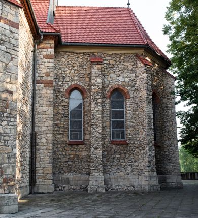 Zdjęcie nr 15: Kościół murowany, orientowany, jednonawowy, trójprzęsłowy, z transeptem i jednoprzęsłowym, węższym i niższym prezbiterium zamkniętym trójbocznie. Między nawą a transeptem po obu stronach znajdują się kruchty, do prezbiterium od strony północnej dostawiona kruchta złożona z dwóch lokalności. Ściany są rozczłonkowane filarami, tworzącymi płytkie arkadowe wnęki. Między nawą a prezbiterium arkada tęczowa. Sklepienie krzyżowe na gurtach spływających na filary. Prezbiterium oświetlają wąskie, prostokątne okna zamknięte ostrołukowo, w nawie i w transepcie znajdują się prostokątne okna prostokątne zamknięte półkoliście, w ścianie frontowej w dolnej części okna prostokątne zamknięte półkoliście, a w górnej podwójne okna prostokątne. Fasada ujęta dwiema narożnymi szkarpami, zwieńczona trójkątnie. Do niej dostawiona kruchta z dwuspadowym daszkiem i wejściem w kształcie prostokąta zamkniętego półkoliście. W kamiennej fasadzie znajdują się dołem okna prostokątne, zamknięte półkoliście, a górą  prostokątne, biforialne okna z nadprożem łukowatym wykonanym z cegły. Szczyt ceglany, podzielony na trzy strefy kamiennym wątkiem, z pięcioma wnękami prostokątnymi, zamkniętymi półkoliście, z wąskimi otworami w dolnej strefie i z owalnym oknem w drugiej. Elewacje boczne nawy ujęte skarpami. Dachy dwuspadowe, kryte dachówką, z wieżyczką na sygnaturkę.         