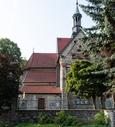 Zdjęcie nr 20: Kościół murowany, orientowany, jednonawowy, trójprzęsłowy, z transeptem i jednoprzęsłowym, węższym i niższym prezbiterium zamkniętym trójbocznie. Między nawą a transeptem po obu stronach znajdują się kruchty, do prezbiterium od strony północnej dostawiona kruchta złożona z dwóch lokalności. Ściany są rozczłonkowane filarami, tworzącymi płytkie arkadowe wnęki. Między nawą a prezbiterium arkada tęczowa. Sklepienie krzyżowe na gurtach spływających na filary. Prezbiterium oświetlają wąskie, prostokątne okna zamknięte ostrołukowo, w nawie i w transepcie znajdują się prostokątne okna prostokątne zamknięte półkoliście, w ścianie frontowej w dolnej części okna prostokątne zamknięte półkoliście, a w górnej podwójne okna prostokątne. Fasada ujęta dwiema narożnymi szkarpami, zwieńczona trójkątnie. Do niej dostawiona kruchta z dwuspadowym daszkiem i wejściem w kształcie prostokąta zamkniętego półkoliście. W kamiennej fasadzie znajdują się dołem okna prostokątne, zamknięte półkoliście, a górą  prostokątne, biforialne okna z nadprożem łukowatym wykonanym z cegły. Szczyt ceglany, podzielony na trzy strefy kamiennym wątkiem, z pięcioma wnękami prostokątnymi, zamkniętymi półkoliście, z wąskimi otworami w dolnej strefie i z owalnym oknem w drugiej. Elewacje boczne nawy ujęte skarpami. Dachy dwuspadowe, kryte dachówką, z wieżyczką na sygnaturkę.         