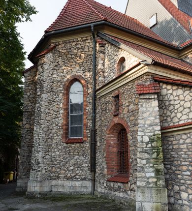 Zdjęcie nr 21: Kościół murowany, orientowany, jednonawowy, trójprzęsłowy, z transeptem i jednoprzęsłowym, węższym i niższym prezbiterium zamkniętym trójbocznie. Między nawą a transeptem po obu stronach znajdują się kruchty, do prezbiterium od strony północnej dostawiona kruchta złożona z dwóch lokalności. Ściany są rozczłonkowane filarami, tworzącymi płytkie arkadowe wnęki. Między nawą a prezbiterium arkada tęczowa. Sklepienie krzyżowe na gurtach spływających na filary. Prezbiterium oświetlają wąskie, prostokątne okna zamknięte ostrołukowo, w nawie i w transepcie znajdują się prostokątne okna prostokątne zamknięte półkoliście, w ścianie frontowej w dolnej części okna prostokątne zamknięte półkoliście, a w górnej podwójne okna prostokątne. Fasada ujęta dwiema narożnymi szkarpami, zwieńczona trójkątnie. Do niej dostawiona kruchta z dwuspadowym daszkiem i wejściem w kształcie prostokąta zamkniętego półkoliście. W kamiennej fasadzie znajdują się dołem okna prostokątne, zamknięte półkoliście, a górą  prostokątne, biforialne okna z nadprożem łukowatym wykonanym z cegły. Szczyt ceglany, podzielony na trzy strefy kamiennym wątkiem, z pięcioma wnękami prostokątnymi, zamkniętymi półkoliście, z wąskimi otworami w dolnej strefie i z owalnym oknem w drugiej. Elewacje boczne nawy ujęte skarpami. Dachy dwuspadowe, kryte dachówką, z wieżyczką na sygnaturkę.         
