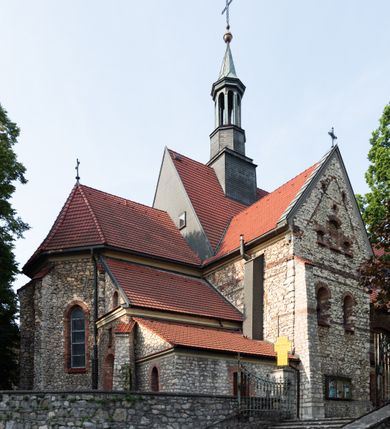 Zdjęcie nr 22: Kościół murowany, orientowany, jednonawowy, trójprzęsłowy, z transeptem i jednoprzęsłowym, węższym i niższym prezbiterium zamkniętym trójbocznie. Między nawą a transeptem po obu stronach znajdują się kruchty, do prezbiterium od strony północnej dostawiona kruchta złożona z dwóch lokalności. Ściany są rozczłonkowane filarami, tworzącymi płytkie arkadowe wnęki. Między nawą a prezbiterium arkada tęczowa. Sklepienie krzyżowe na gurtach spływających na filary. Prezbiterium oświetlają wąskie, prostokątne okna zamknięte ostrołukowo, w nawie i w transepcie znajdują się prostokątne okna prostokątne zamknięte półkoliście, w ścianie frontowej w dolnej części okna prostokątne zamknięte półkoliście, a w górnej podwójne okna prostokątne. Fasada ujęta dwiema narożnymi szkarpami, zwieńczona trójkątnie. Do niej dostawiona kruchta z dwuspadowym daszkiem i wejściem w kształcie prostokąta zamkniętego półkoliście. W kamiennej fasadzie znajdują się dołem okna prostokątne, zamknięte półkoliście, a górą  prostokątne, biforialne okna z nadprożem łukowatym wykonanym z cegły. Szczyt ceglany, podzielony na trzy strefy kamiennym wątkiem, z pięcioma wnękami prostokątnymi, zamkniętymi półkoliście, z wąskimi otworami w dolnej strefie i z owalnym oknem w drugiej. Elewacje boczne nawy ujęte skarpami. Dachy dwuspadowe, kryte dachówką, z wieżyczką na sygnaturkę.         