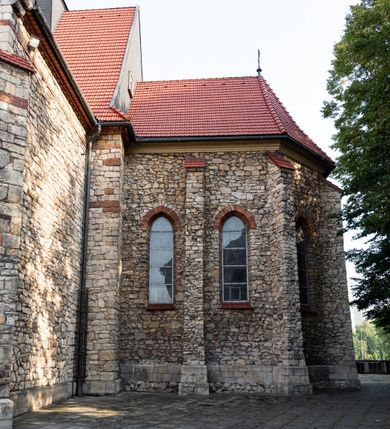 Zdjęcie nr 32: Kościół murowany, orientowany, jednonawowy, trójprzęsłowy, z transeptem i jednoprzęsłowym, węższym i niższym prezbiterium zamkniętym trójbocznie. Między nawą a transeptem po obu stronach znajdują się kruchty, do prezbiterium od strony północnej dostawiona kruchta złożona z dwóch lokalności. Ściany są rozczłonkowane filarami, tworzącymi płytkie arkadowe wnęki. Między nawą a prezbiterium arkada tęczowa. Sklepienie krzyżowe na gurtach spływających na filary. Prezbiterium oświetlają wąskie, prostokątne okna zamknięte ostrołukowo, w nawie i w transepcie znajdują się prostokątne okna prostokątne zamknięte półkoliście, w ścianie frontowej w dolnej części okna prostokątne zamknięte półkoliście, a w górnej podwójne okna prostokątne. Fasada ujęta dwiema narożnymi szkarpami, zwieńczona trójkątnie. Do niej dostawiona kruchta z dwuspadowym daszkiem i wejściem w kształcie prostokąta zamkniętego półkoliście. W kamiennej fasadzie znajdują się dołem okna prostokątne, zamknięte półkoliście, a górą  prostokątne, biforialne okna z nadprożem łukowatym wykonanym z cegły. Szczyt ceglany, podzielony na trzy strefy kamiennym wątkiem, z pięcioma wnękami prostokątnymi, zamkniętymi półkoliście, z wąskimi otworami w dolnej strefie i z owalnym oknem w drugiej. Elewacje boczne nawy ujęte skarpami. Dachy dwuspadowe, kryte dachówką, z wieżyczką na sygnaturkę.         